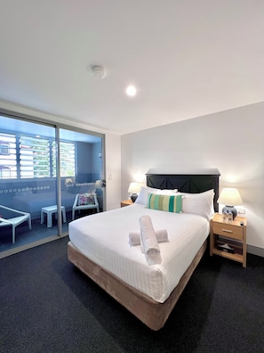 Habitación de hotel con una cama grande, dos mesitas de noche, una puerta corrediza de vidrio que da a un balcón y vistas al exterior.