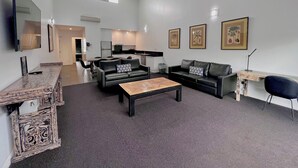 A modern living room with a sofa, coffee table, and a dining area with chairs. There is a kitchen in the background, a TV mounted on the wall, and framed pictures on the wall.