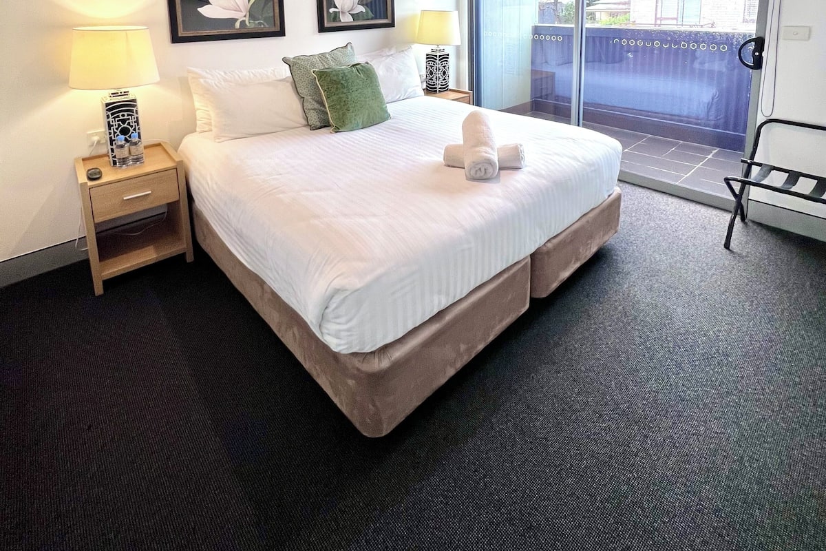 A hotel room with a large bed, two nightstands with lamps, a view of the outside through a sliding door, and two framed floral paintings on the wall.
