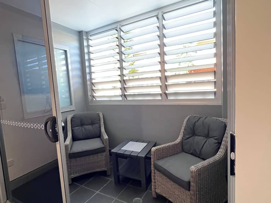 Una habitación con puerta de vidrio corrediza, dos sillas de mimbre, una mesita y una ventana con persianas.