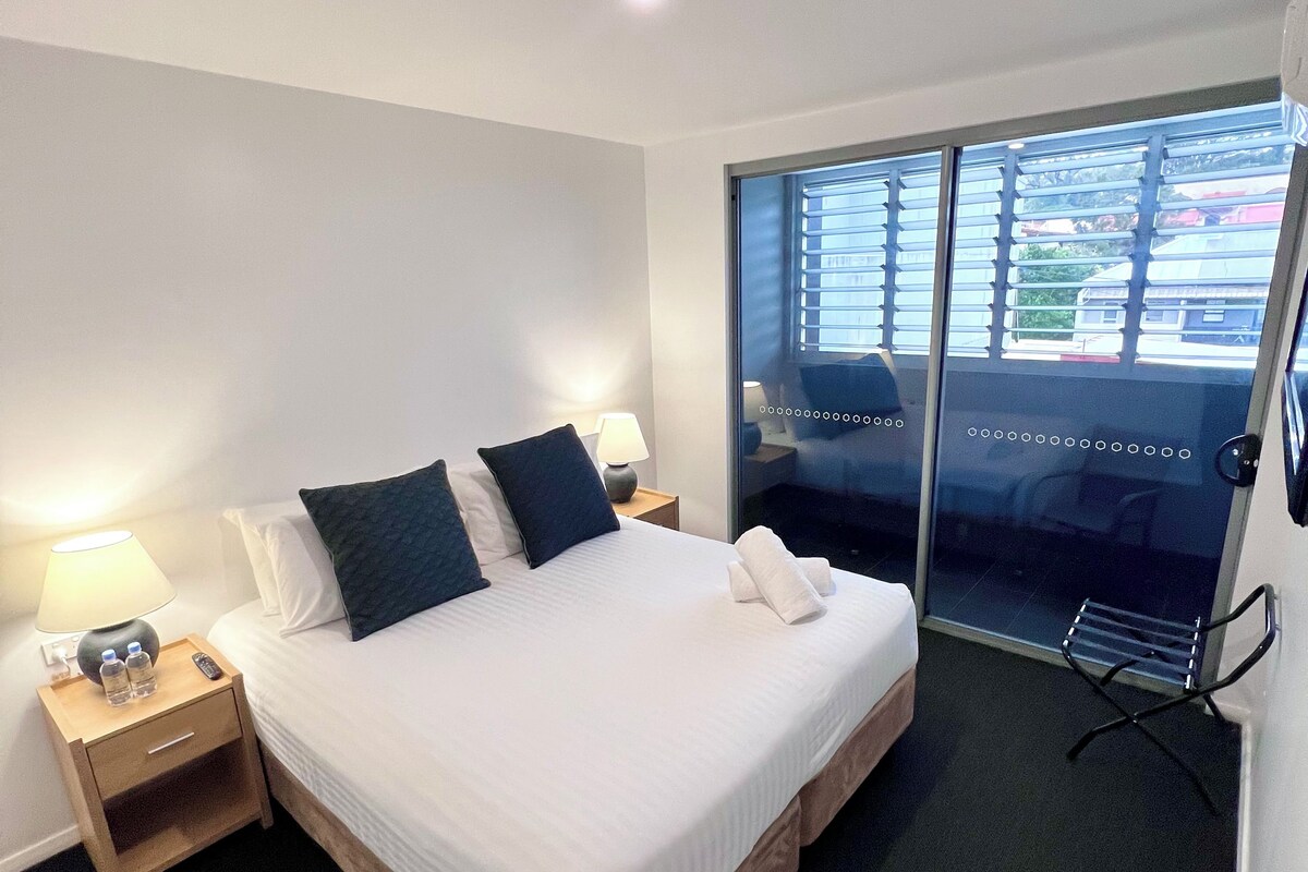 A hotel room with a bed, bedside tables, a lamp, a towel, and a view through a glass door.