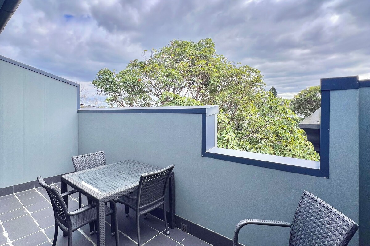 A balcony with a table and chairs, a view of trees, and a cloudy sky.