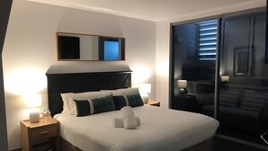 A hotel room with a large bed, two bedside tables with lamps, a mirror above the bed, and a sliding glass door leading to a balcony.