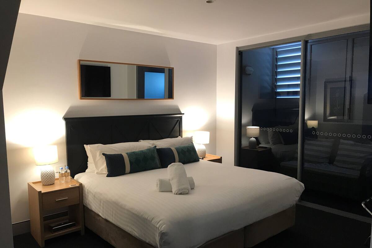 A hotel room with a large bed, two bedside tables with lamps, a mirror above the bed, and a sliding glass door leading to a balcony.