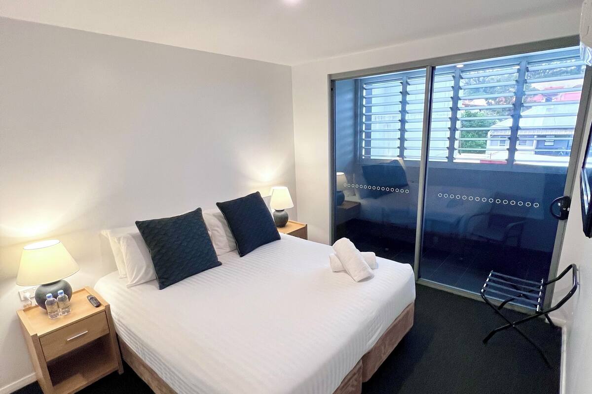 A hotel room with a bed, bedside tables, a lamp, a towel, and a view through a glass door.