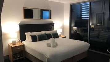 A hotel room with a large bed, two bedside tables with lamps, a mirror above the bed, and a sliding glass door leading to a balcony.