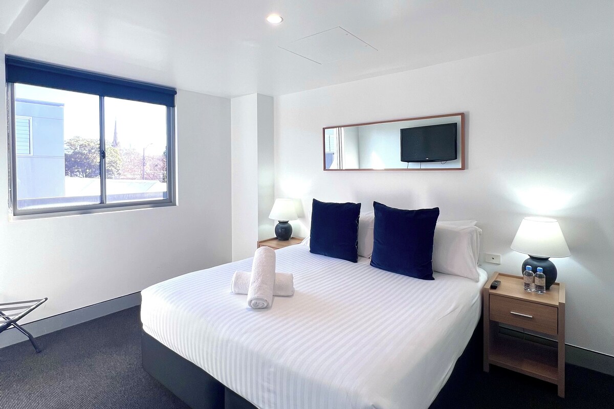 A hotel room with a large bed, a bedside table with a lamp, a TV mounted on the wall, and a window with a view of buildings.