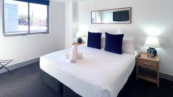 A hotel room with a large bed, a bedside table with a lamp, a TV mounted on the wall, and a window with a view of buildings.