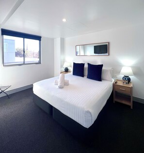 Habitación de hotel con una cama grande, una mesita de noche con lámpara, un televisor montado en la pared y una ventana con vistas a edificios.