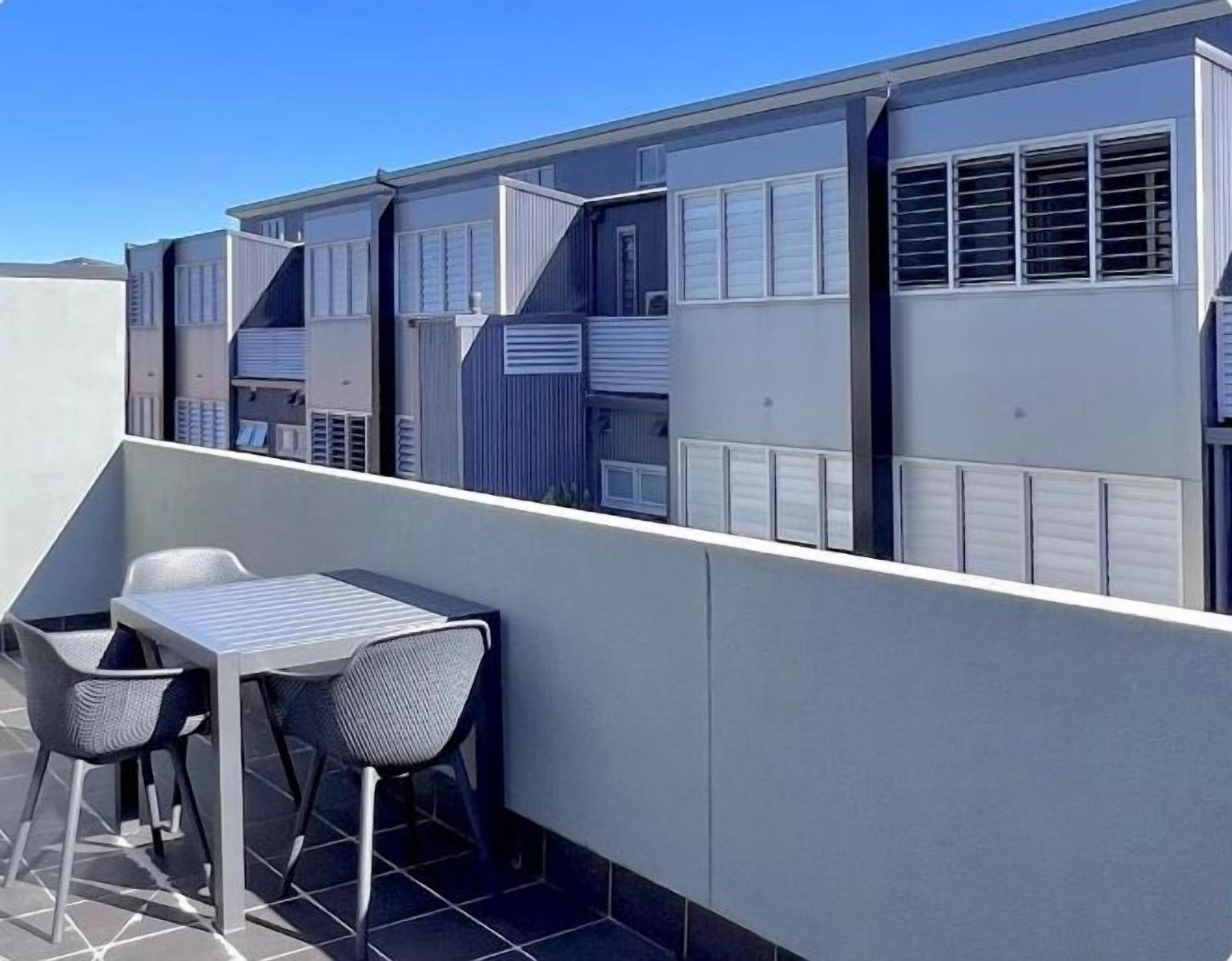Una terrazza sul tetto con un tavolo e due sedie, affacciata su una fila di edifici con ampie finestre e bocchette di aerazione.
