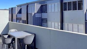 Una terrazza sul tetto con un tavolo e due sedie, affacciata su una fila di edifici con ampie finestre e bocchette di aerazione.