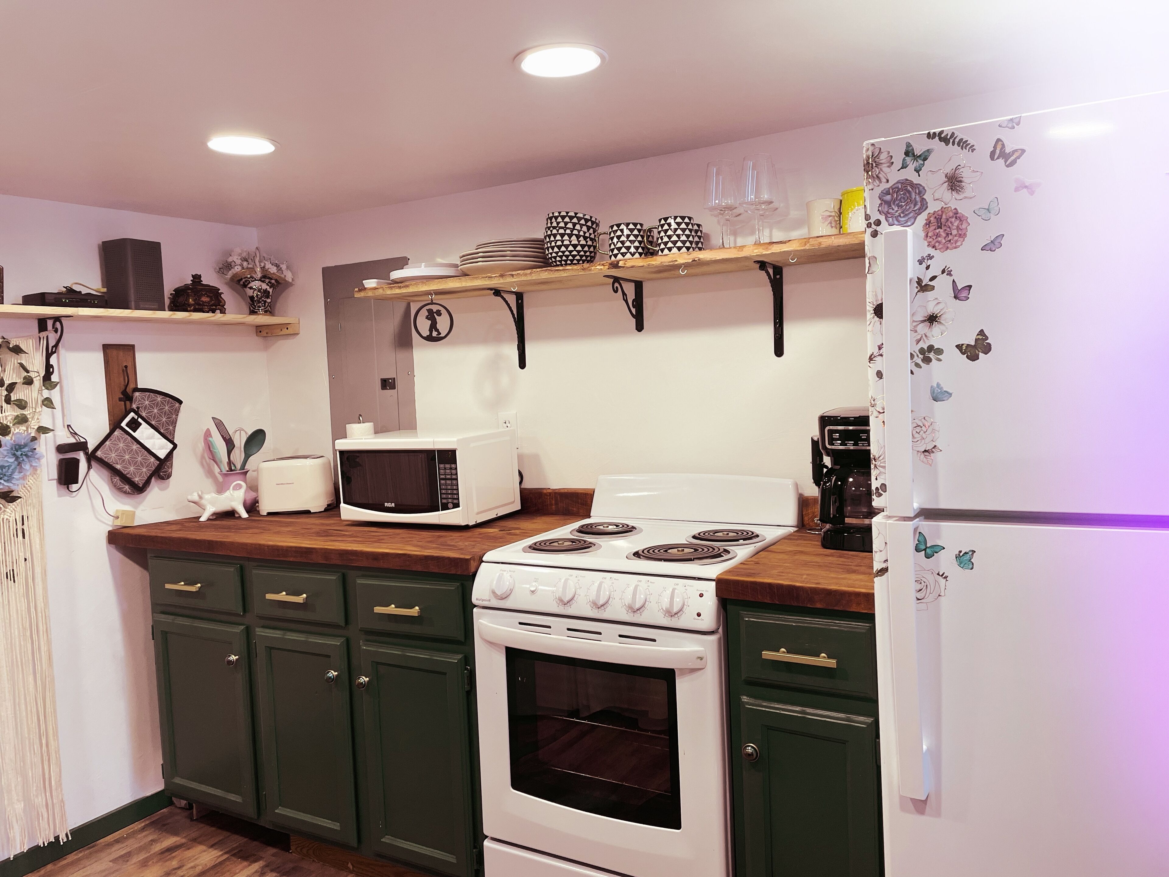 A kitchen with a microwave, stove, oven, and a refrigerator. There are shelves above the counter holding dishes and glasses.