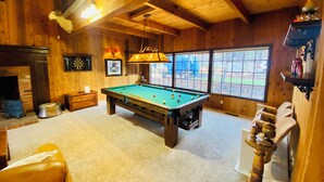 A pool table in a wooden room with a fireplace, dartboard, and a window with a view. - Charming 4-bedroom Cabin in Peaceful Lake Arrowhead (Lake Arrowhead)