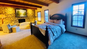A bedroom with a stone wall, a bed with a wooden headboard, a stone fireplace, a window, and a view of trees. - Charming 4-bedroom Cabin in Peaceful Lake Arrowhead (Lake Arrowhead)