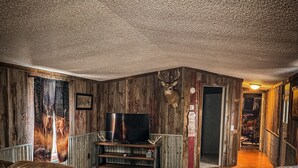 A rustic living room with wooden walls, a mounted deer head, leather sofas, a TV, and a wooden floor. - 3-bedroom cabin located in Steuben County home of 101 lakes! (Angola)
