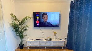 A living room with a television mounted on the wall displaying a show, a white console table with various items, a potted plant, and a round coffee table with snacks. - Home Away From Home! Lovely House Near M1 (Nottingham)