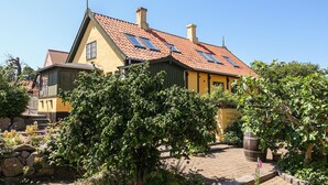 Una casa tradizionale con tetto spiovente, diversi camini e un giardino con varie piante e una botte grande.