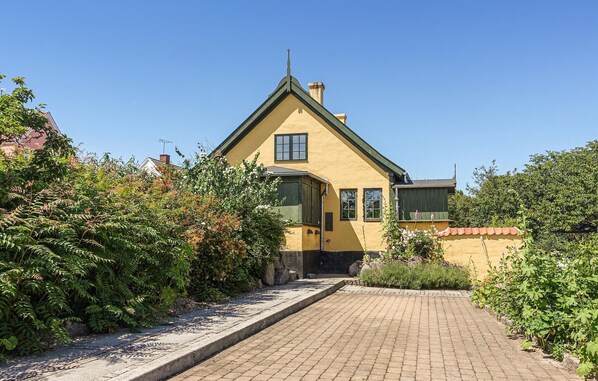 A yellow house with a green roof, surrounded by dense greenery and a paved pathway leading to the entrance.