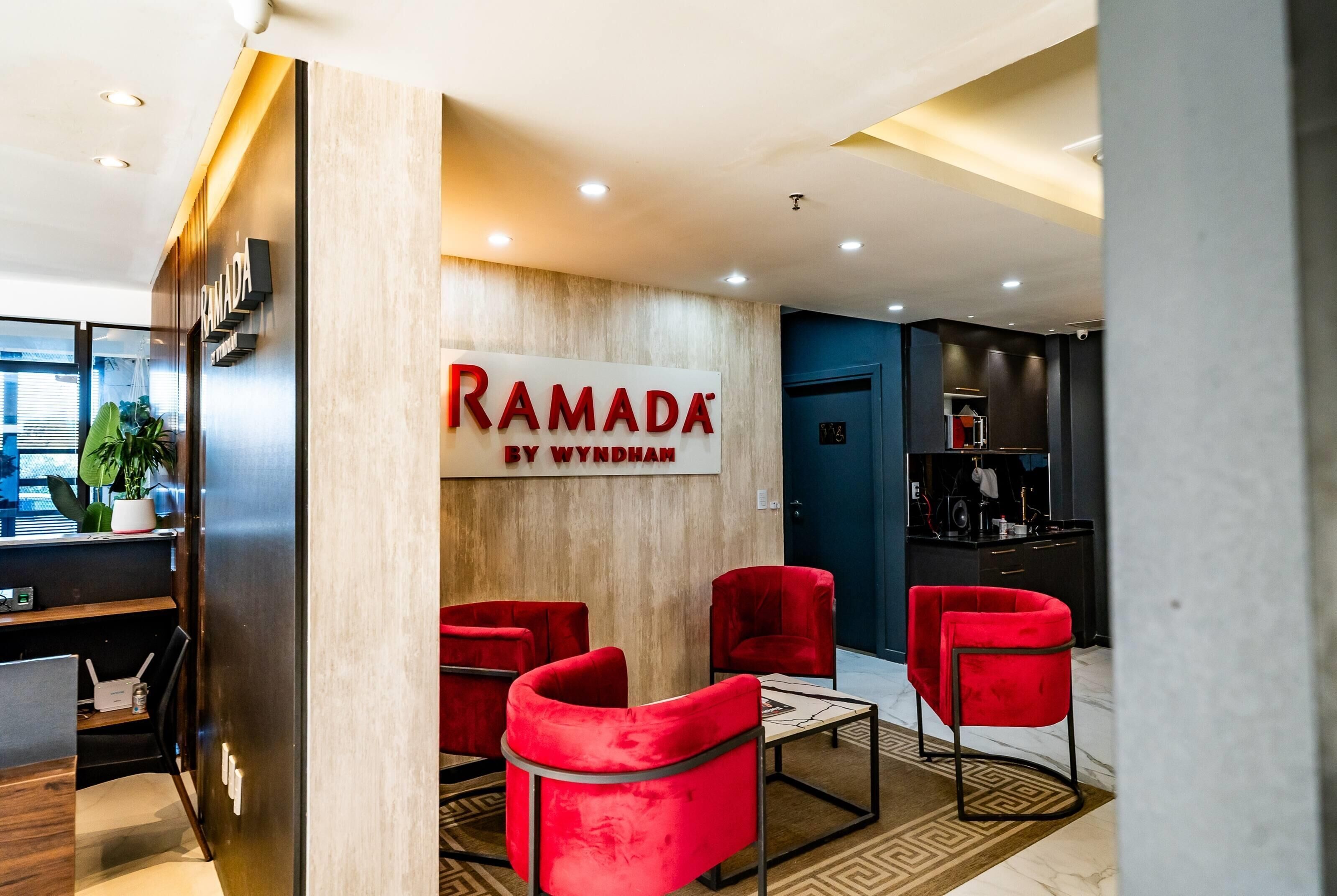 A modern reception area with a wooden wall, a sign reading "RAMADA BY WYNDHAM", and red chairs.