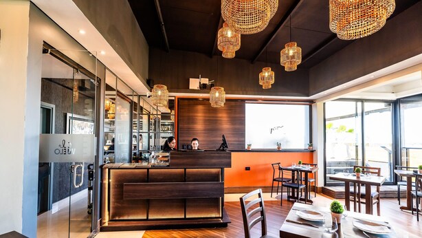 A modern restaurant interior with a wooden counter, glass partitions, and hanging wicker chandeliers.