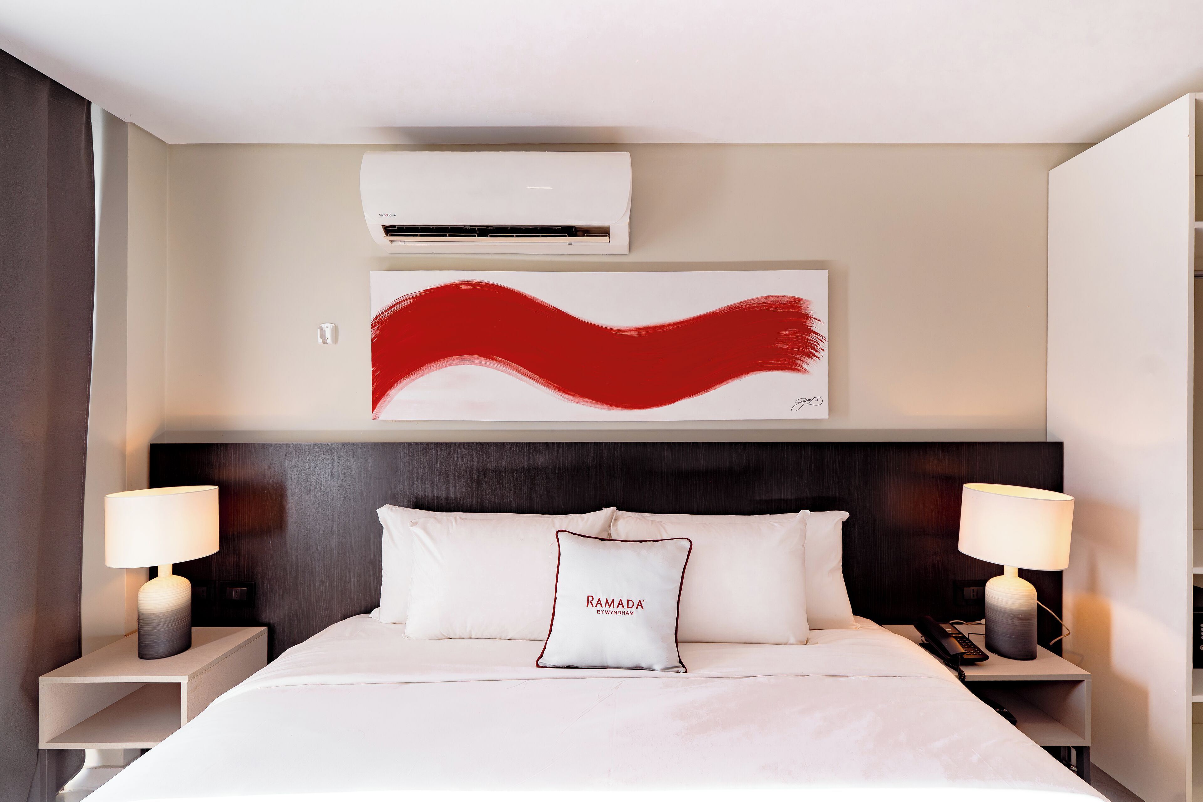 Habitación de hotel con cama, dos lámparas de noche, un cuadro abstracto grande, un equipo de aire acondicionado y una almohada con un nombre bordado.