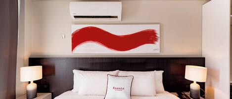 Habitación de hotel con cama, dos lámparas de noche, un cuadro abstracto grande, un equipo de aire acondicionado y una almohada con un nombre bordado.