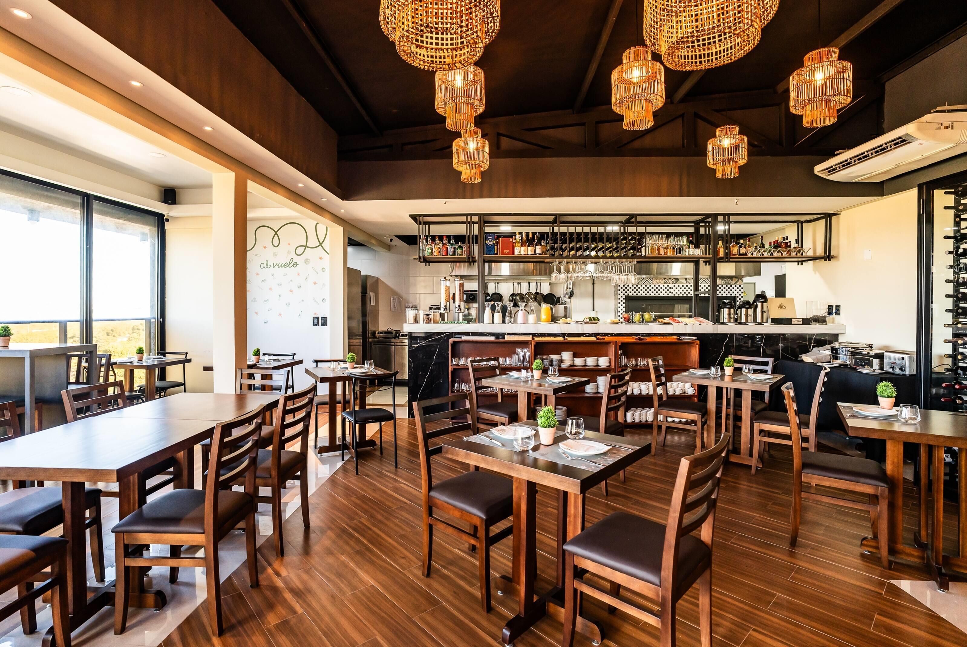 Interior de un restaurante con varias mesas y sillas para comer, una zona de bar con estantes llenos de botellas, y una cocina al fondo.