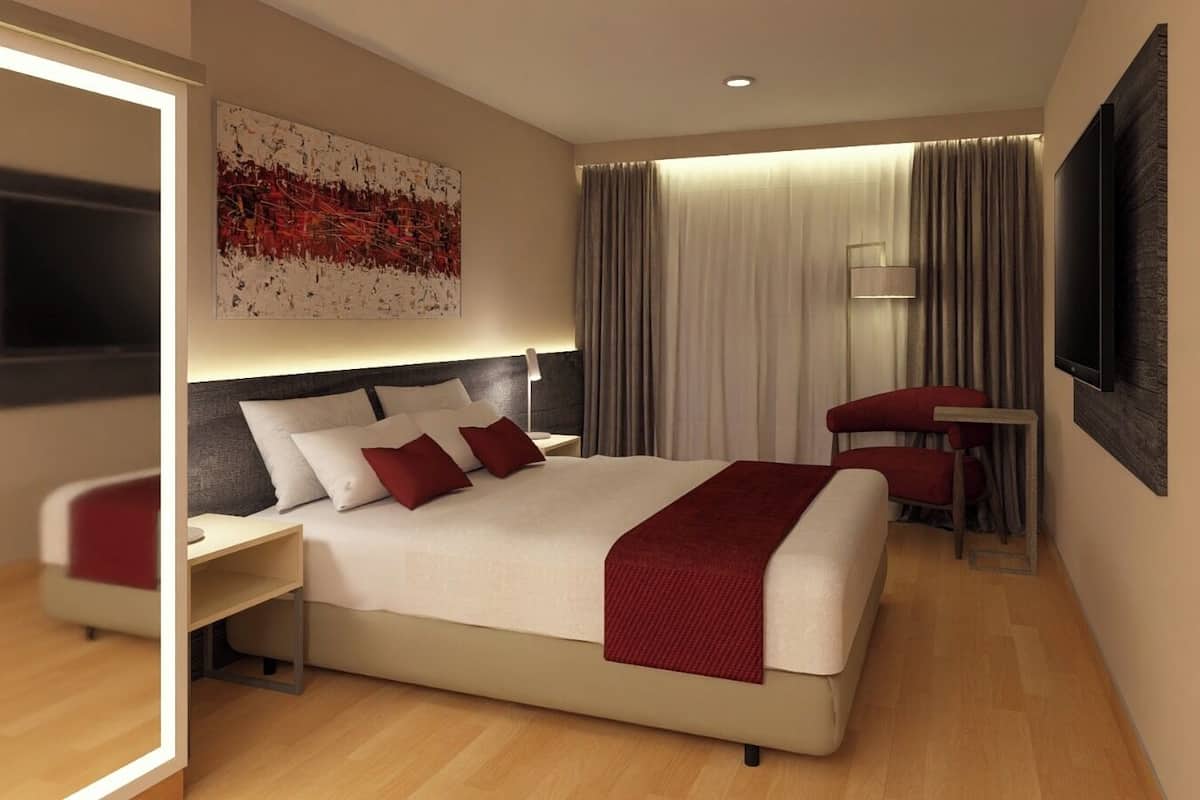 Quarto de hotel moderno com uma cama grande, mesa de cabeceira, poltrona, televisão e uma grande pintura abstrata na parede.