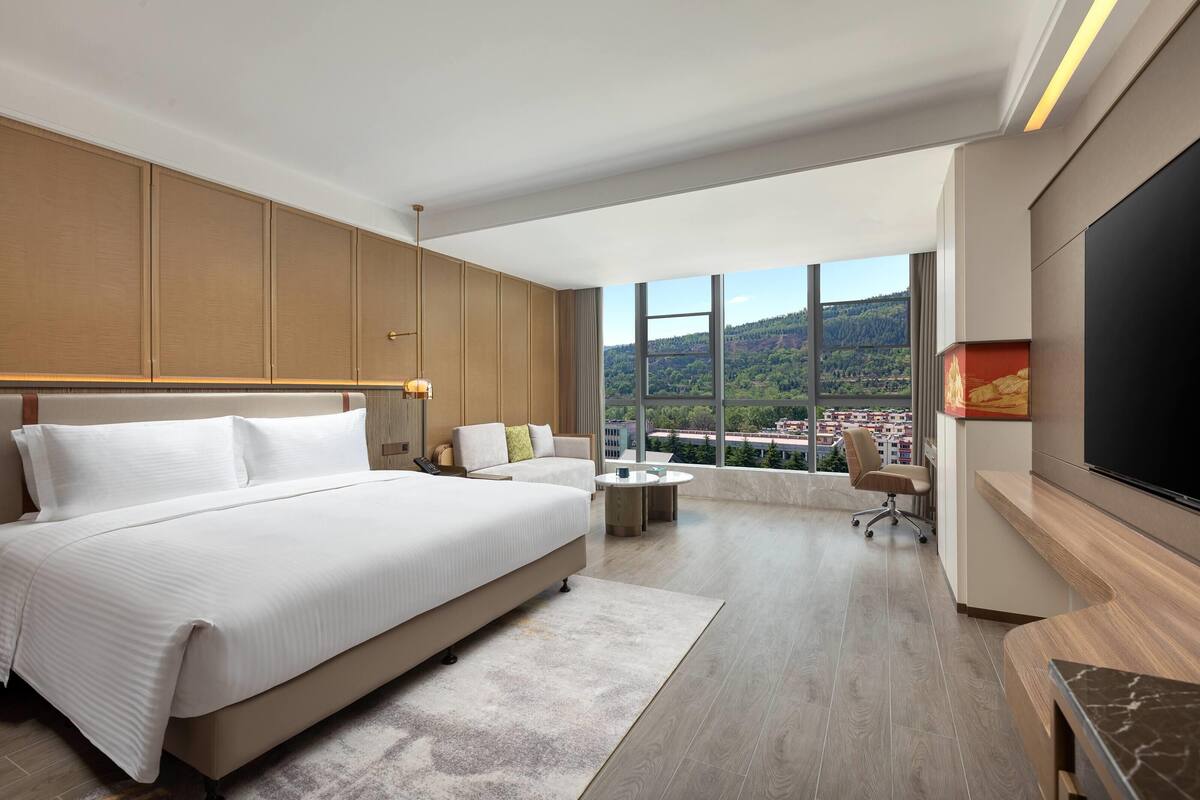 Une chambre d’hôtel moderne dotée d’un grand lit, d’un bureau, d’une chaise et d’une télévision. La chambre dispose de grandes fenêtres offrant une vue sur la ville.