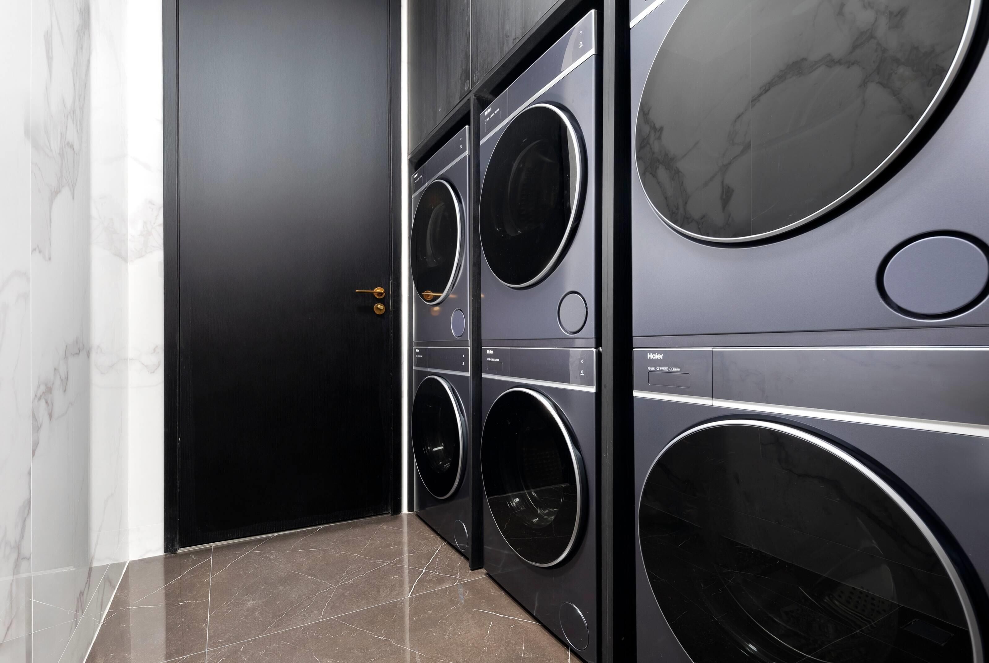 A laundry room with four stacked front-loading washing machines, a black door, and a marble-tiled floor.