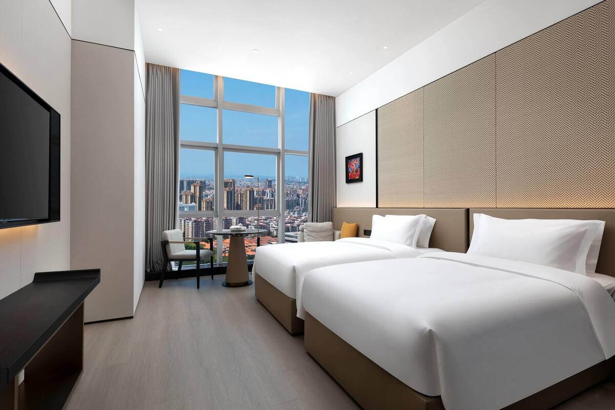 A hotel room with a large window offering a city view, a bed, a chair, a small table, and a TV.