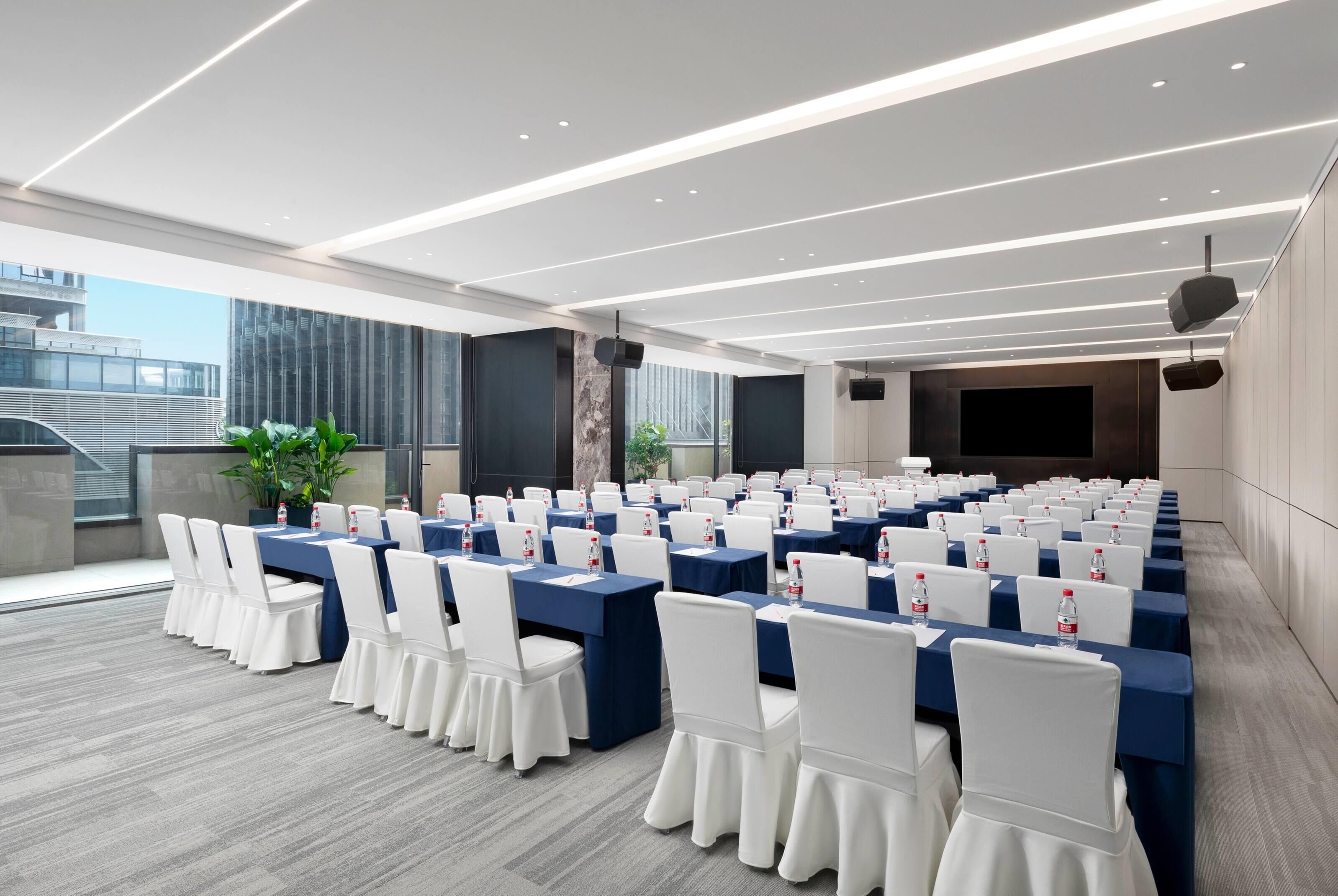 A conference room with rows of tables and chairs, large windows, and a modern ceiling design.