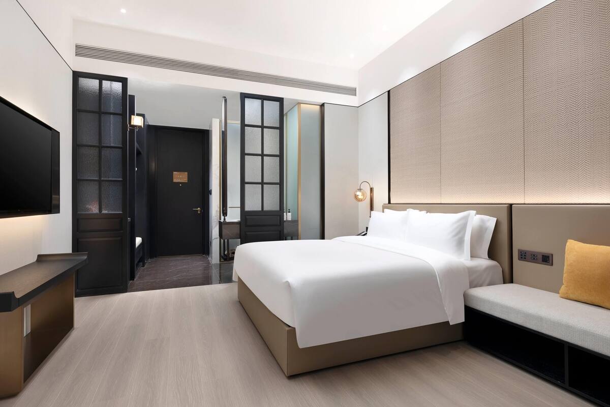 A modern hotel room with a large bed, a seating area, a desk, and a television.