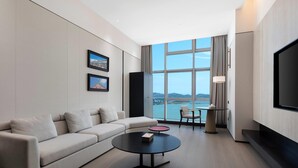 Una habitación de hotel moderna con un ventanal grande, un sofá, una mesa baja, un televisor y una mesita de noche con lámpara.