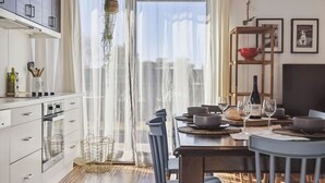 Une cuisine moderne avec des meubles blancs, un coin repas doté d’une table en bois dressée pour deux personnes, et une vue à travers des rideaux légers.