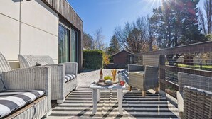 Une terrasse extérieure avec des meubles en osier, une table garnie de nourriture et de boissons, et une vue sur des arbres et un bâtiment en arrière-plan.