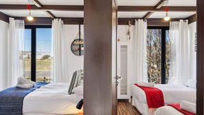 Une chambre d’hôtel avec deux lits, une grande fenêtre avec des rideaux, une étagère fixée au mur ornée d’objets décoratifs et un plancher en bois.