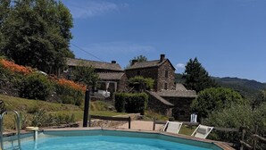 A swimming pool with a wooden deck, surrounded by stone buildings and lush greenery. - Cosy gîte with view, jacuzzi & pool - heart of the Cévennes, hiking, relaxation. (Ventalon en Cévennes)