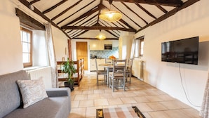 A cozy living room with a sofa, a dining table with chairs, a kitchen in the background, and a TV mounted on the wall. - Cosy gîte with view, jacuzzi & pool - heart of the Cévennes, hiking, relaxation. (Ventalon en Cévennes)