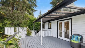 A wooden deck with a glass enclosure, white exterior walls, and a wicker chair with a cushion.