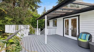 A wooden deck with a glass enclosure, white exterior walls, and a wicker chair with a cushion.