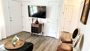 A living room with a black leather ottoman, a television on a stand, and two white chairs with brown leather seats. - Newnan Gem (Newnan)