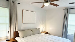 A bedroom with a bed, a nightstand, a bedside lamp, a ceiling fan, and a window with curtains. - Newnan Gem (Newnan)