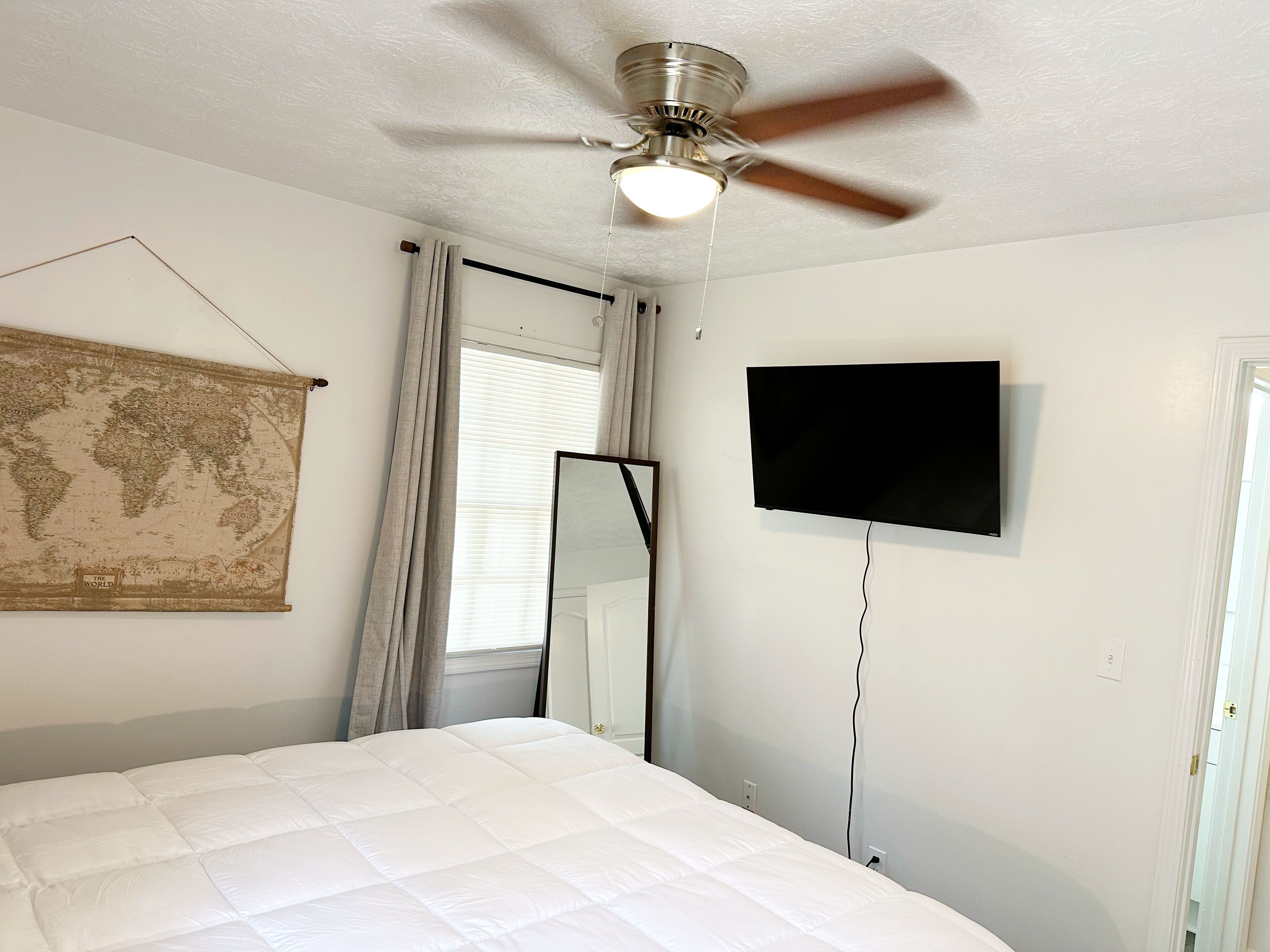 Une chambre à coucher avec un lit, un ventilateur de plafond, une carte du monde, un téléviseur et une fenêtre avec des rideaux.