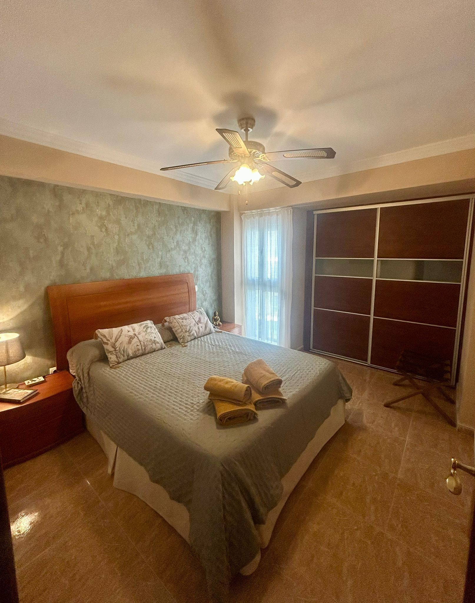 Ein Schlafzimmer mit Bett, Nachttisch, Deckenventilator, Kleiderschrank und einem Fenster mit Vorhängen.