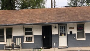 A single-story building with a dark gray and white exterior, a black door, and a small window with a 'OPEN' sign. There are two chairs outside and a small garden area with plants.