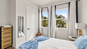Une chambre à coucher avec un lit, une commode, un miroir, une fenêtre avec des rideaux et une vue sur des palmiers.