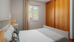 A bedroom with a bed, a nightstand, a lamp, a window with curtains, and a wooden wardrobe. - With outdoor space - Moradia Felicidade Rocha PXO (Lombas)