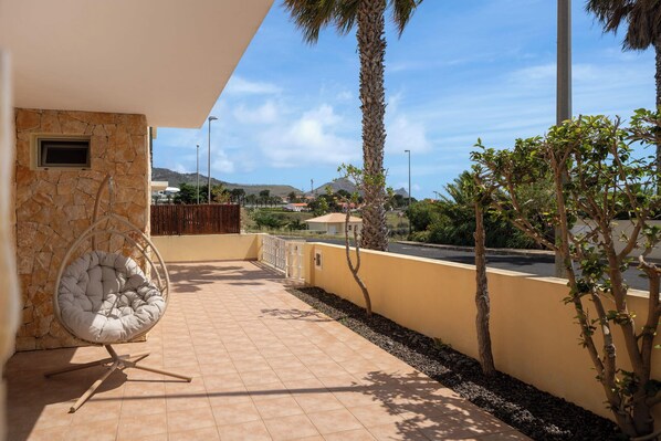 A balcony with a stone wall, a cushioned chair, and a view of a mountain and houses in the distance.