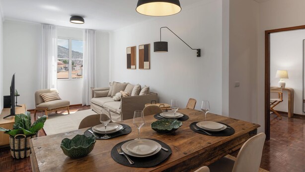 A modern living room with a wooden dining table set for four, a sofa, a TV, and a view of the city. - Quebra Costas I – A Refuge with Panoramic Views (Funchal)